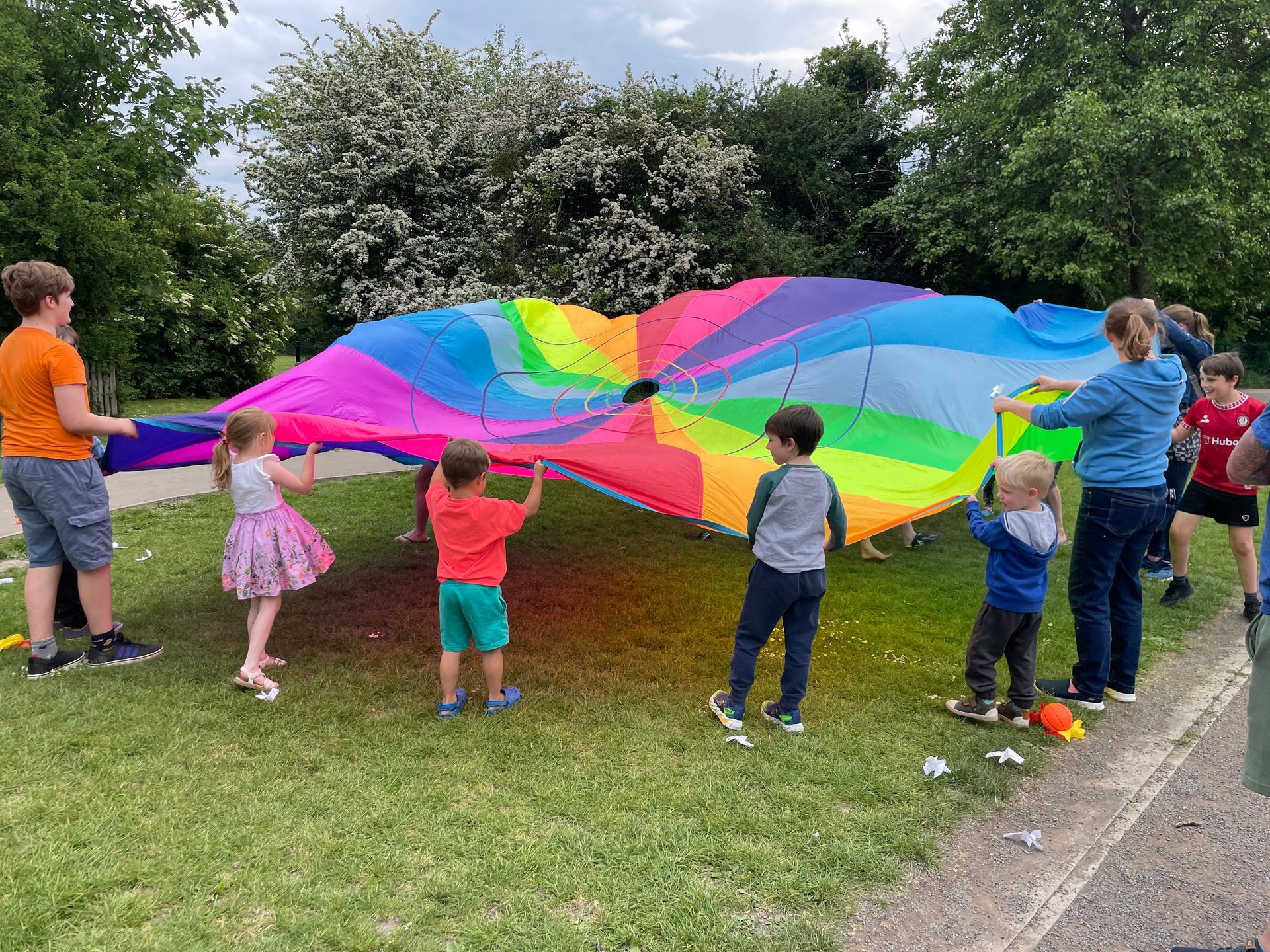 Children and adults playing with a rainbow parachute on a grassy field.