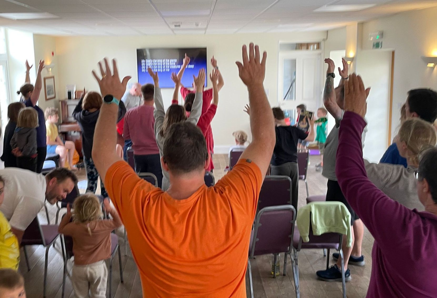 People with arms raised in a room. Some are near chairs. A screen is visible in the background.