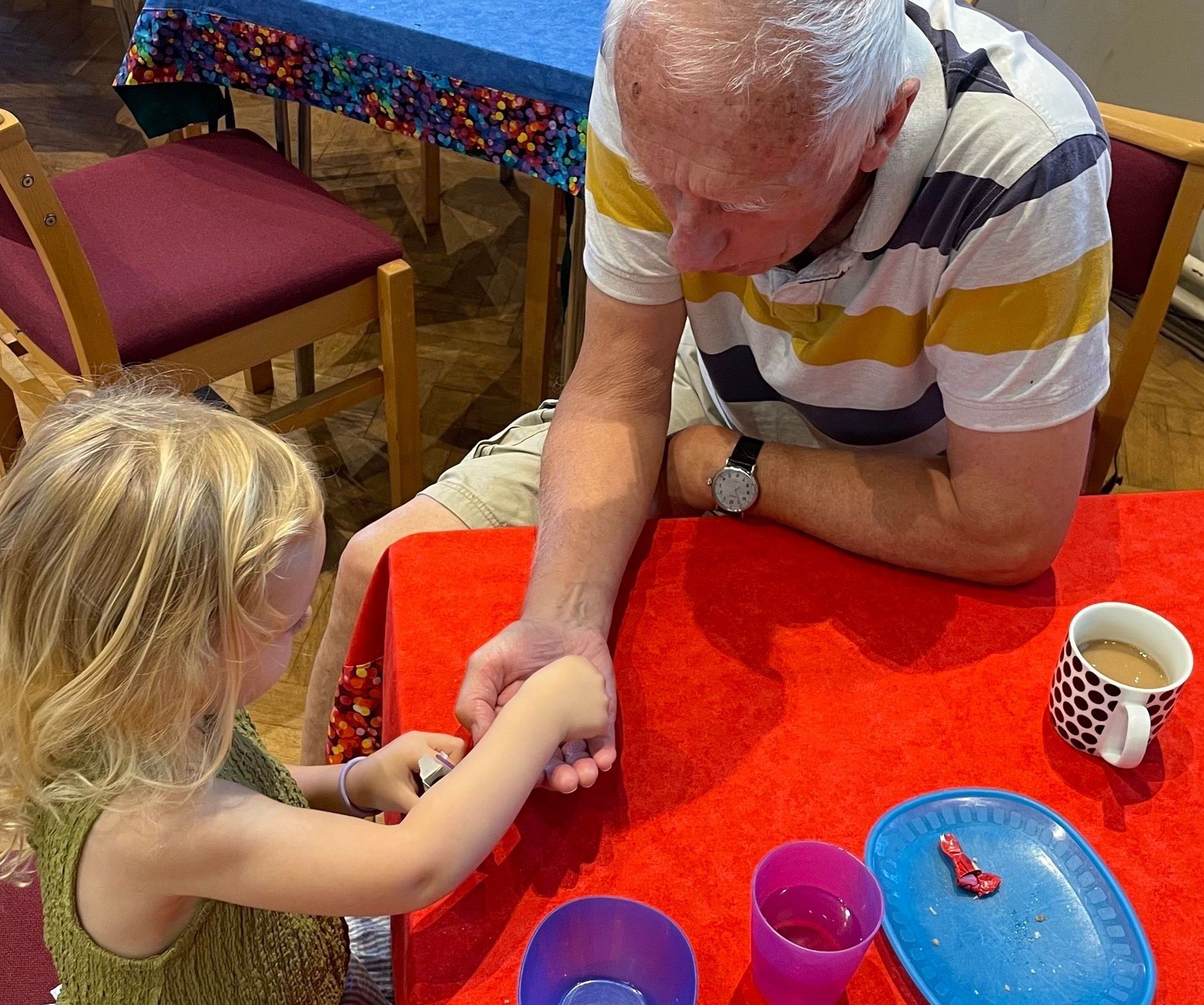 Child and man at a table, child holding man's wrist. Red table cloth, coffee cup, and plates visible.