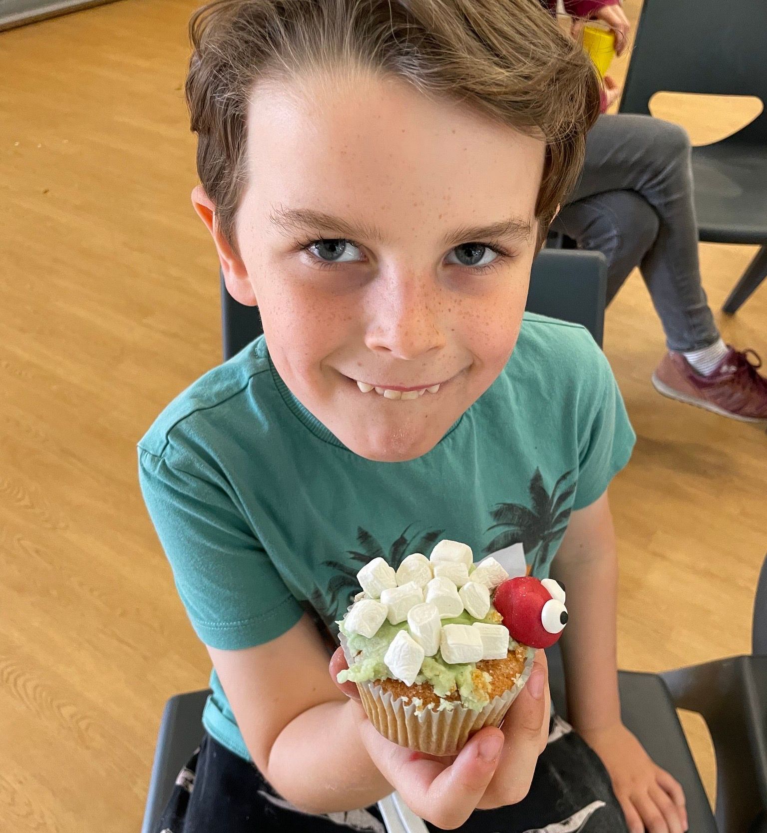 Boy holding a cupcake with marshmallow topping and a decorative candy insect, smiling indoors.