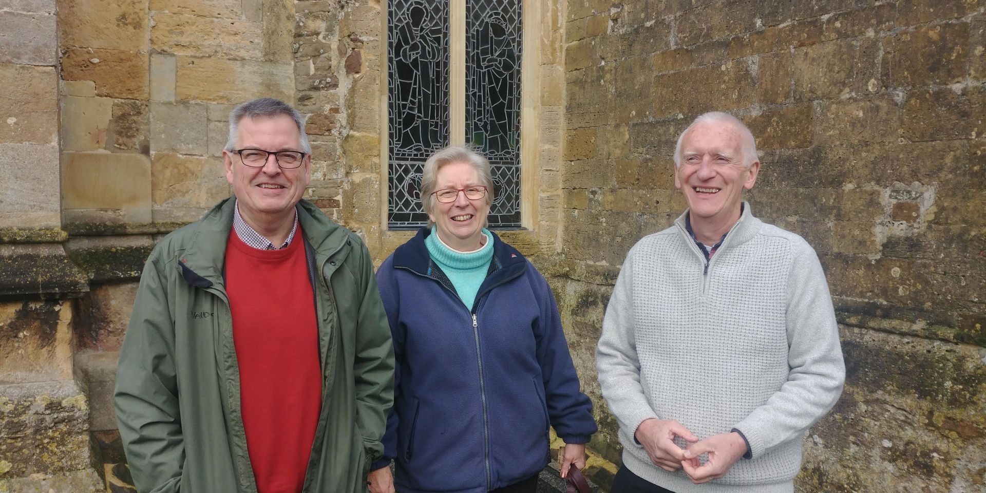 Three people standing in front of a stone building with a window. They are smiling.