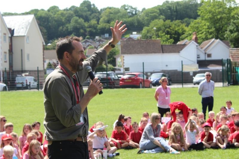 Man speaking into a microphone outdoors, gesturing to a seated group of children in red shirts.
