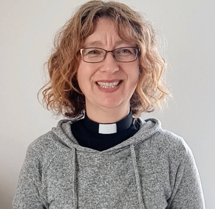 Woman with curly hair and glasses wearing a clergy collar and grey hoodie, smiling.