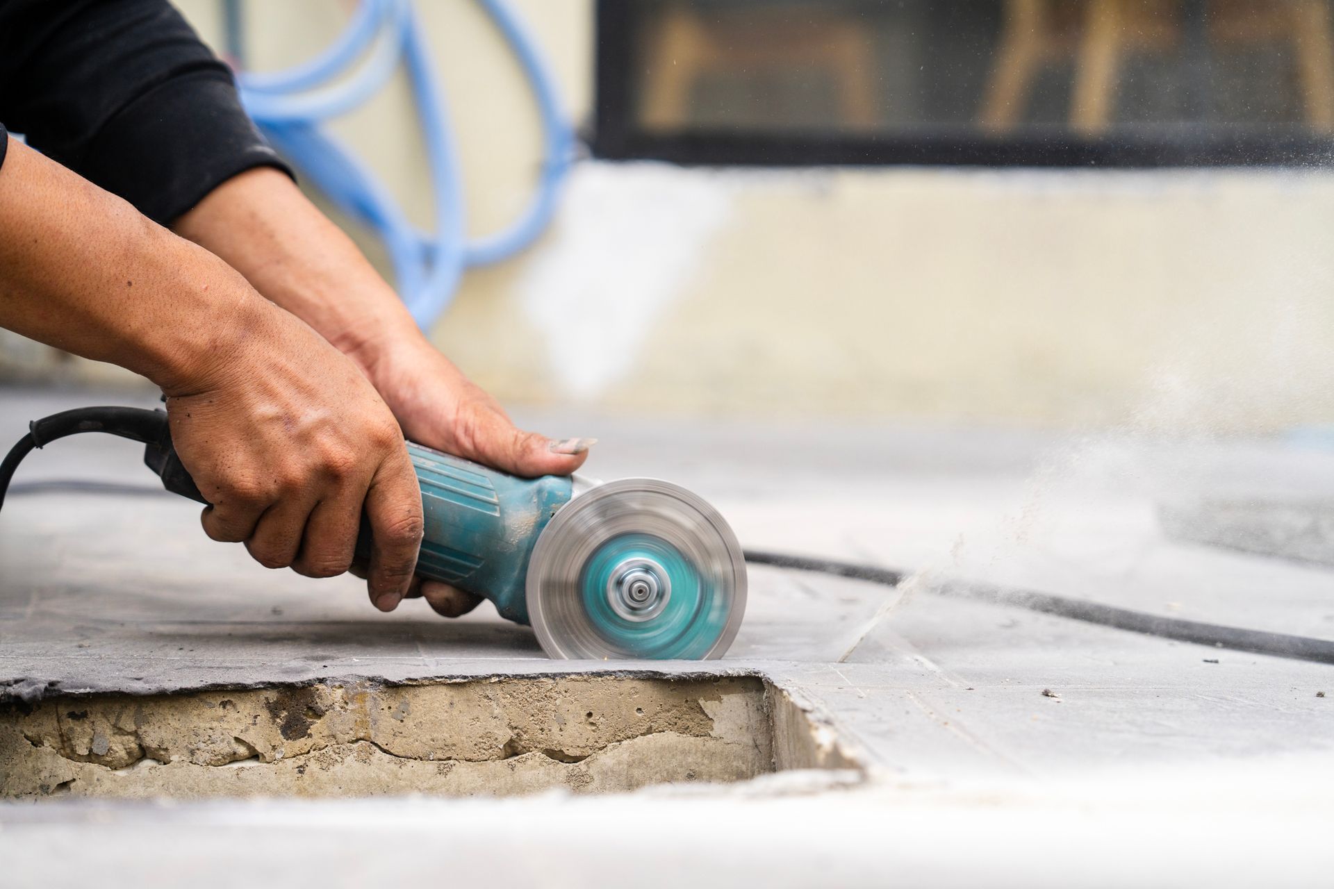 A pair of hands uses a blue concrete grinder on a slab of concrete on the floor.