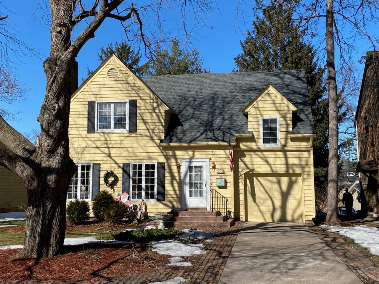 A yellow house with black shutters is surrounded by snow and trees.