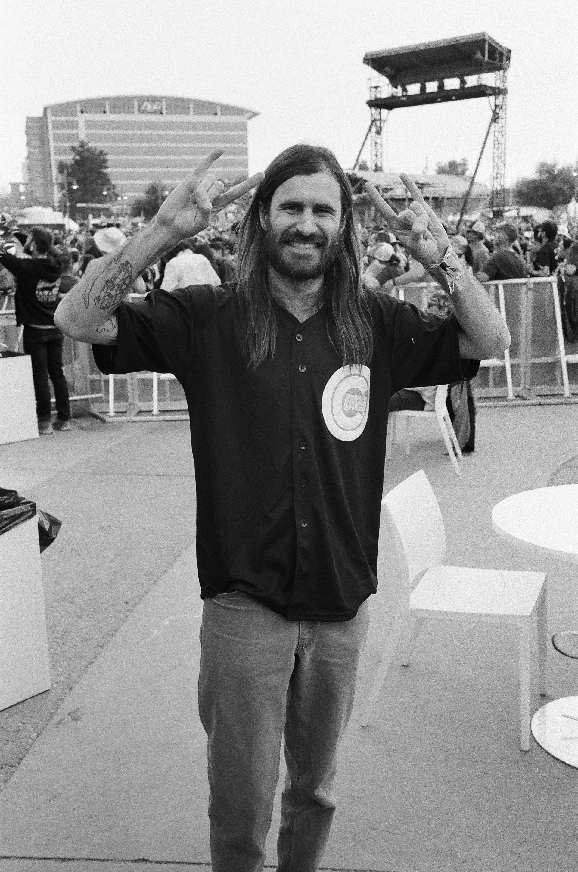 A man with long hair and a beard is standing in front of a crowd giving the peace sign.