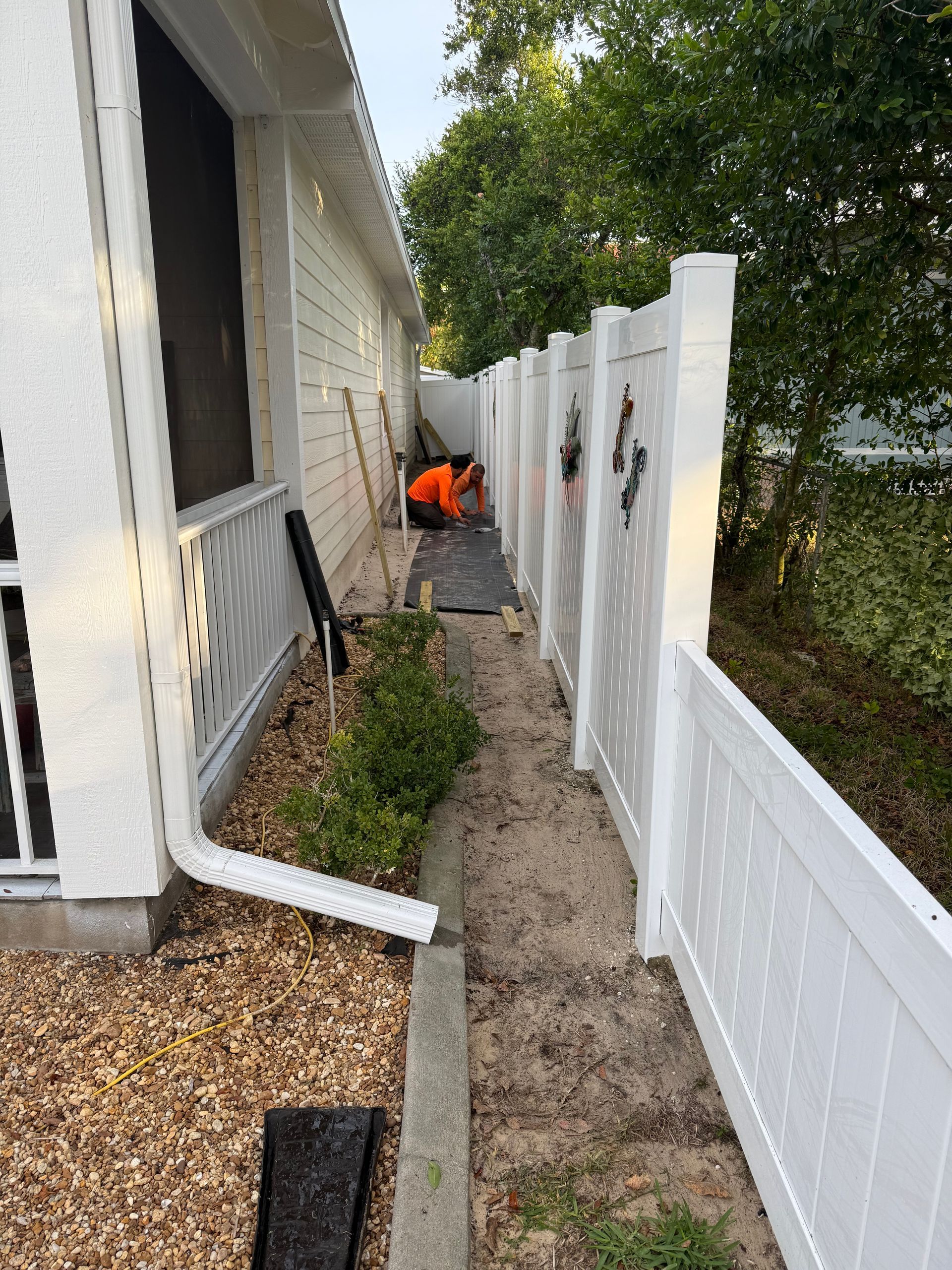 Side view of a house and white fence. A person in orange works on the ground. A narrow dirt path is between them.