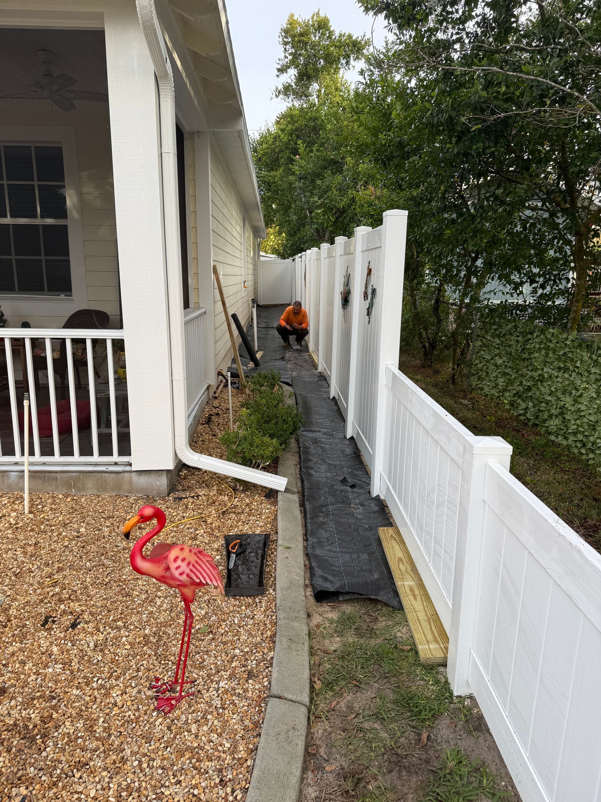 A person works in a narrow yard between a house and white fence. Red flamingo yard art in gravel.