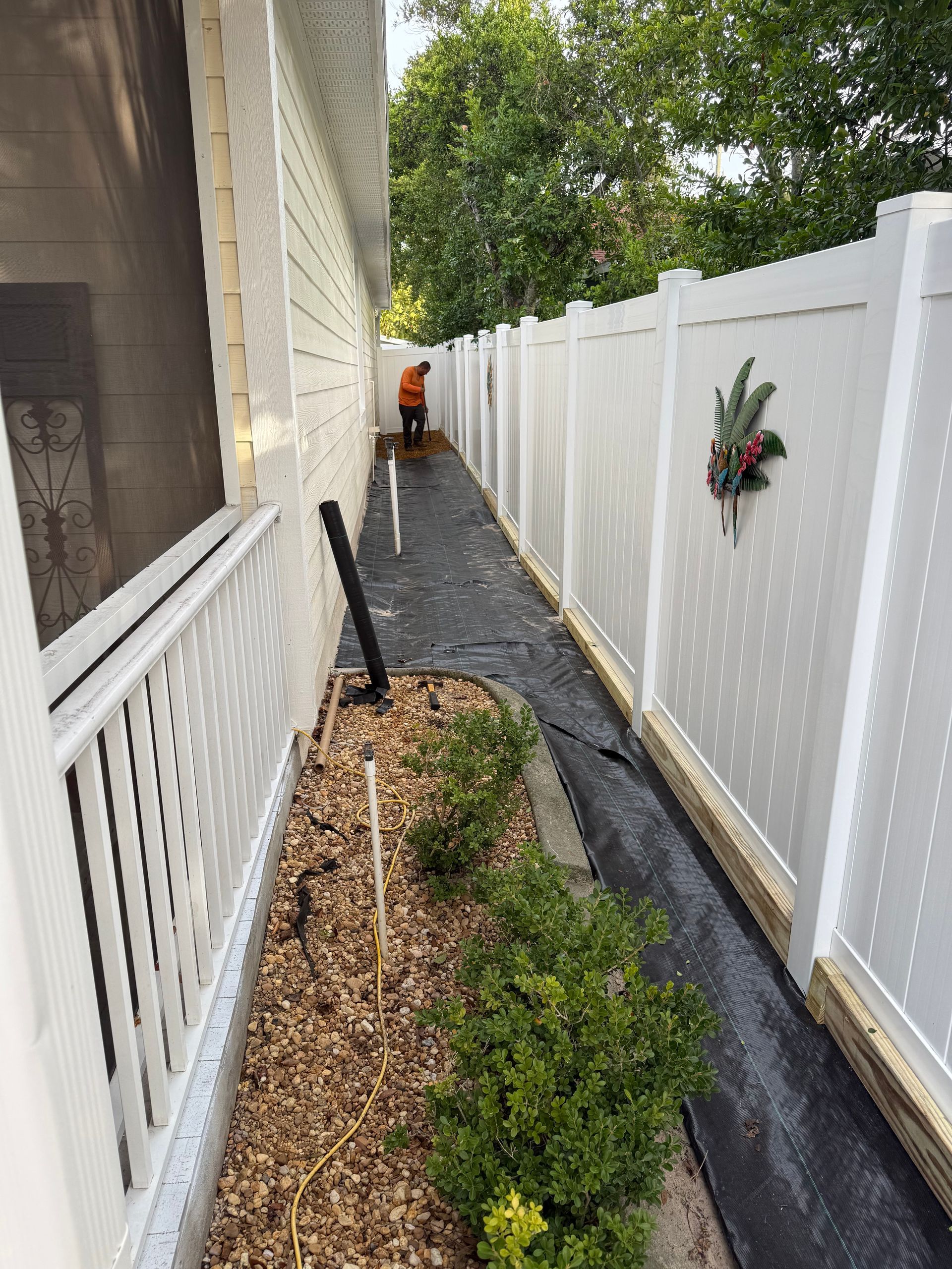 Side yard with a white fence, landscaping, and a person working in the distance.