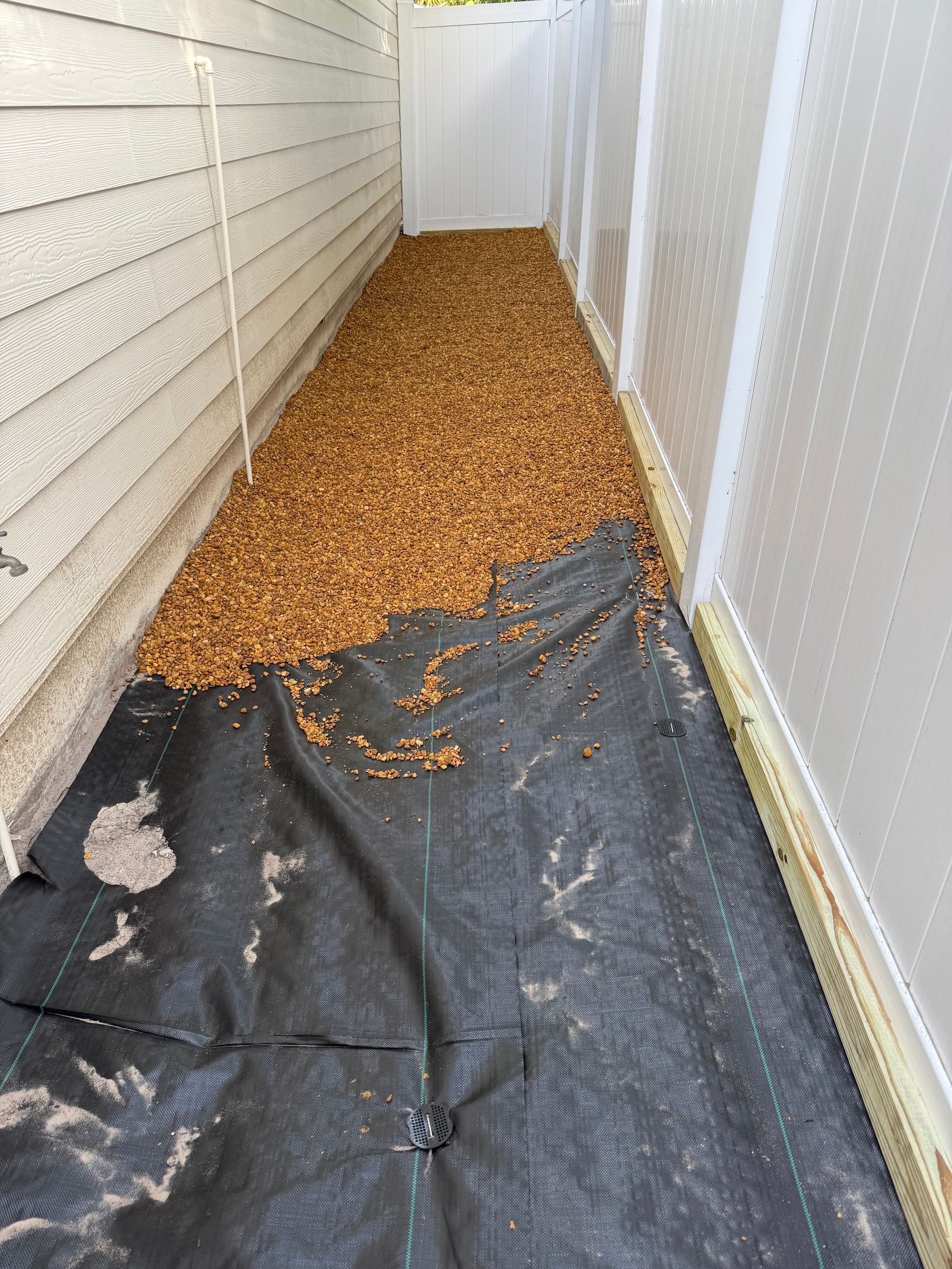 A narrow walkway between a white building and a fence. Black landscape fabric is covered with brown gravel.