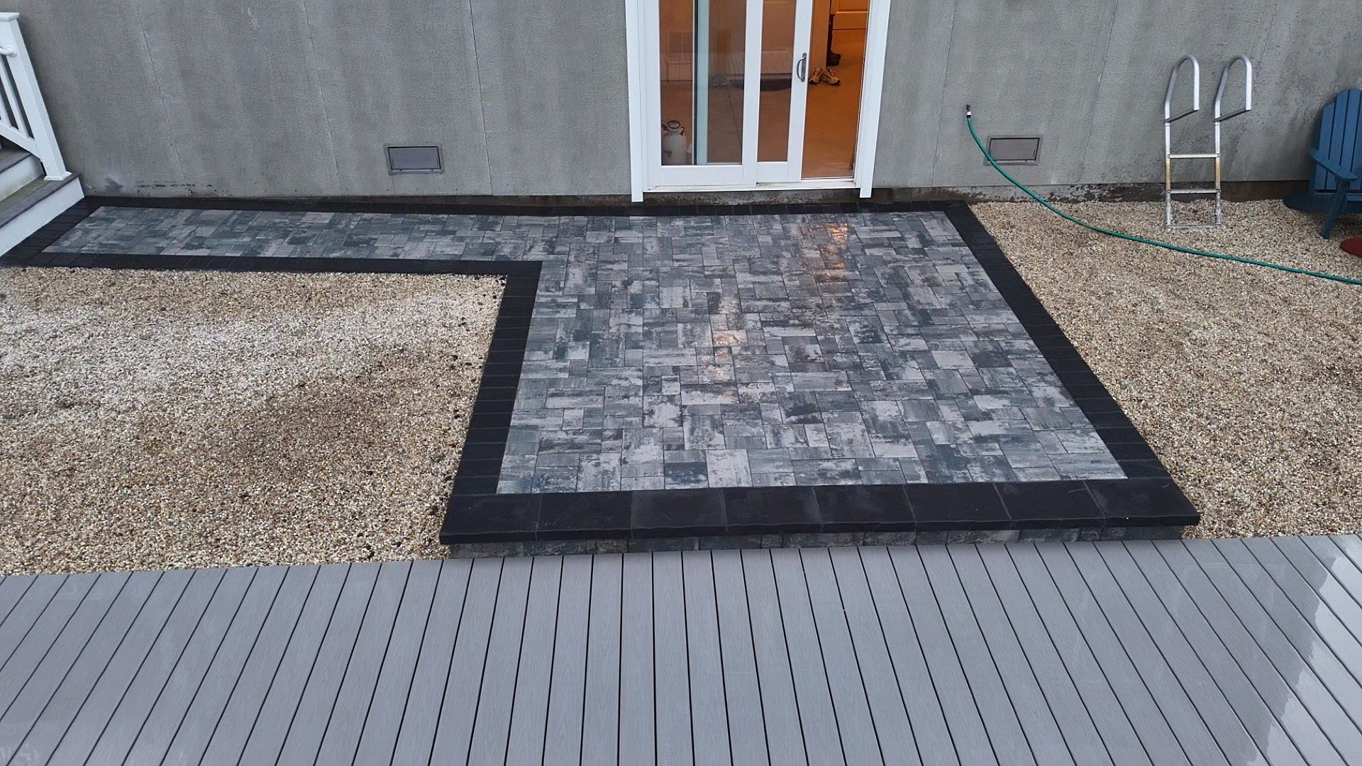 Patio with stone pavers and black border adjacent to gravel and a deck. Sliding glass door visible.