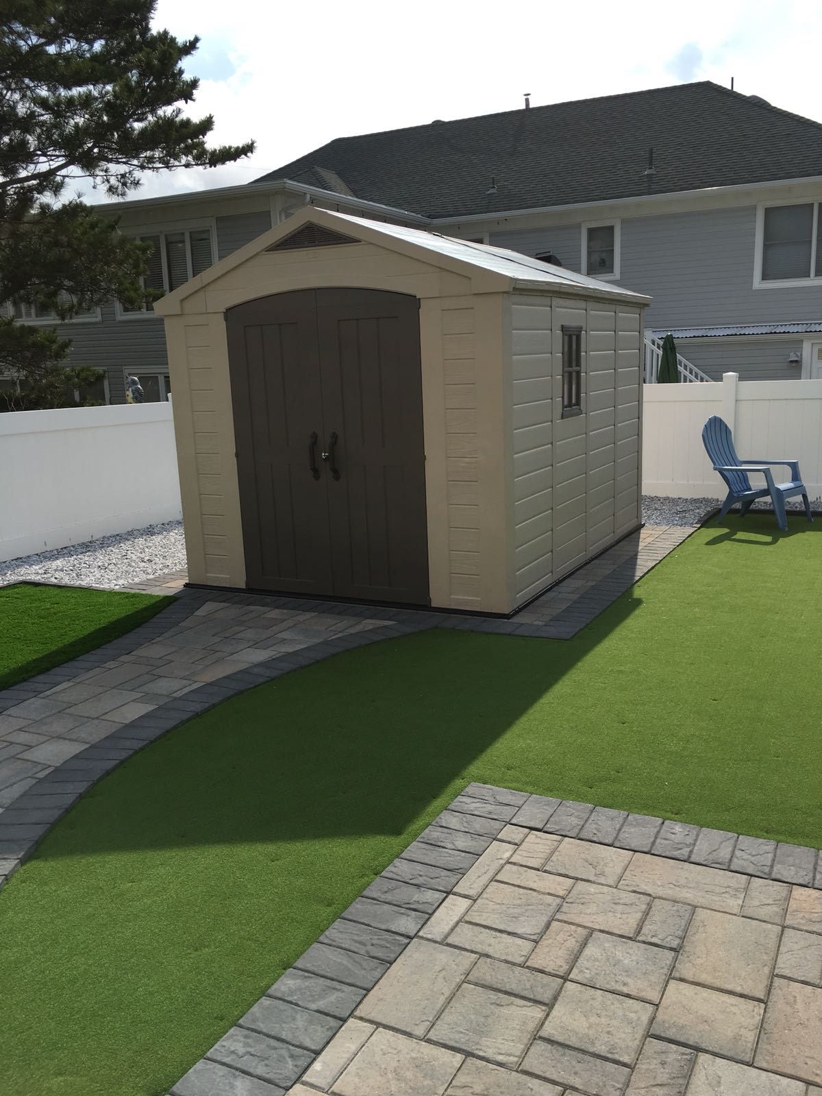 Tan and brown shed with double doors, set on a brick pathway in a backyard with green turf and a blue chair.