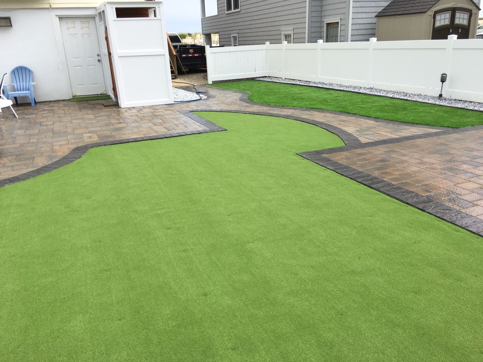 Backyard with green artificial turf surrounded by patterned concrete and white fence.
