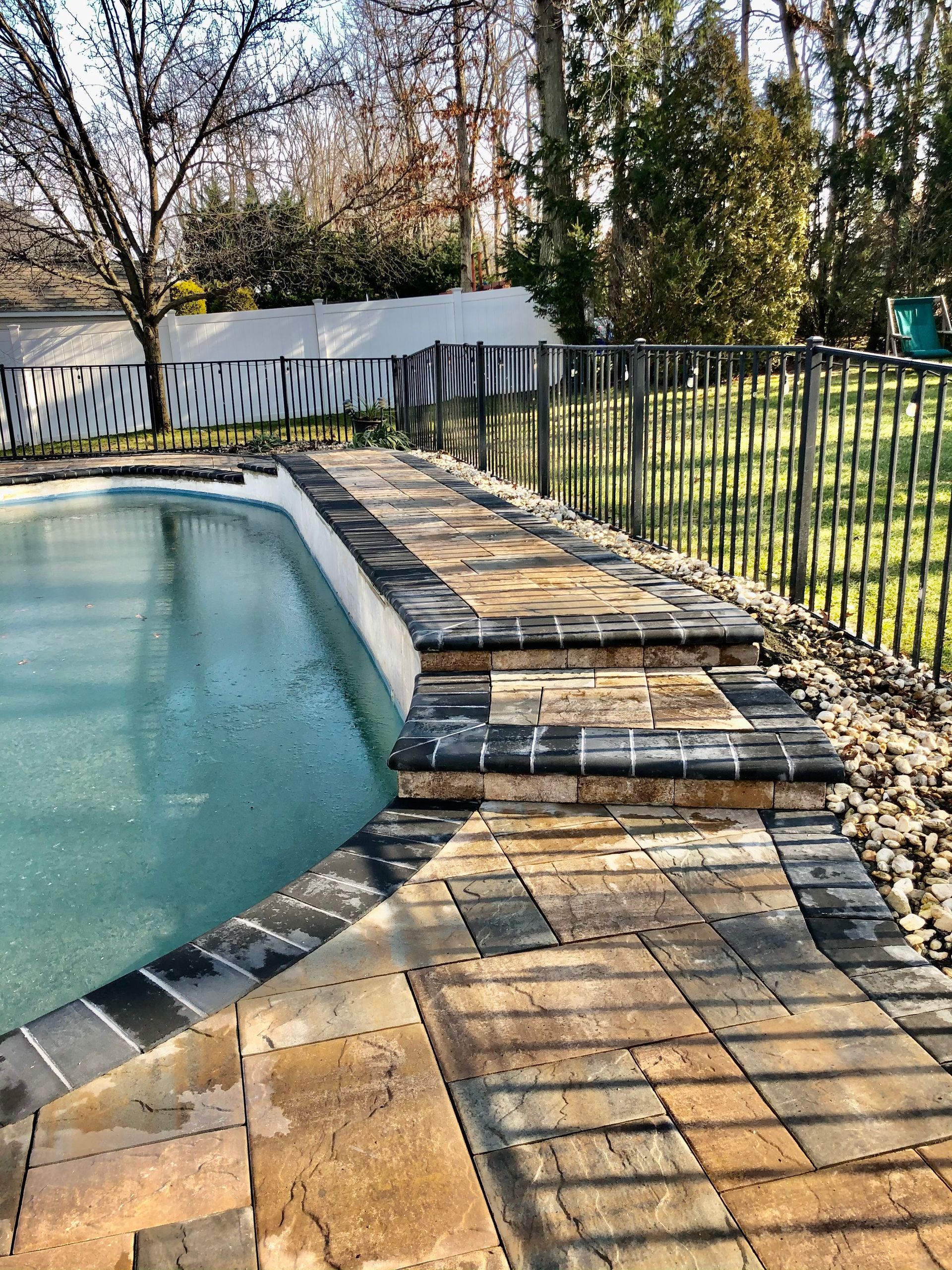 Poolside patio with steps, brick pavers, black metal fence, and a partially visible lawn.