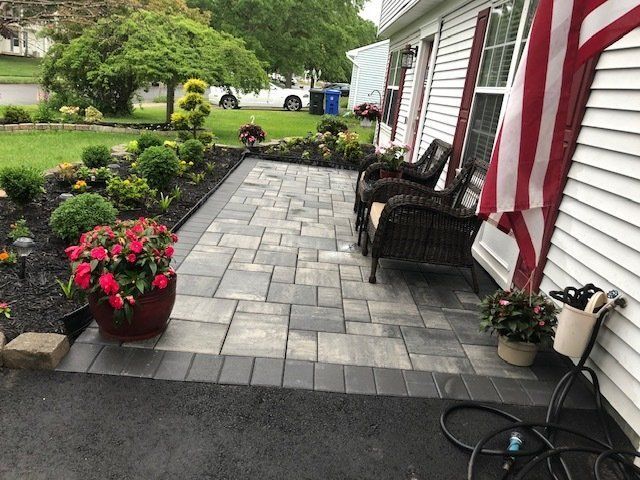 A brick patio with two wicker chairs near a house with an American flag and landscaping.