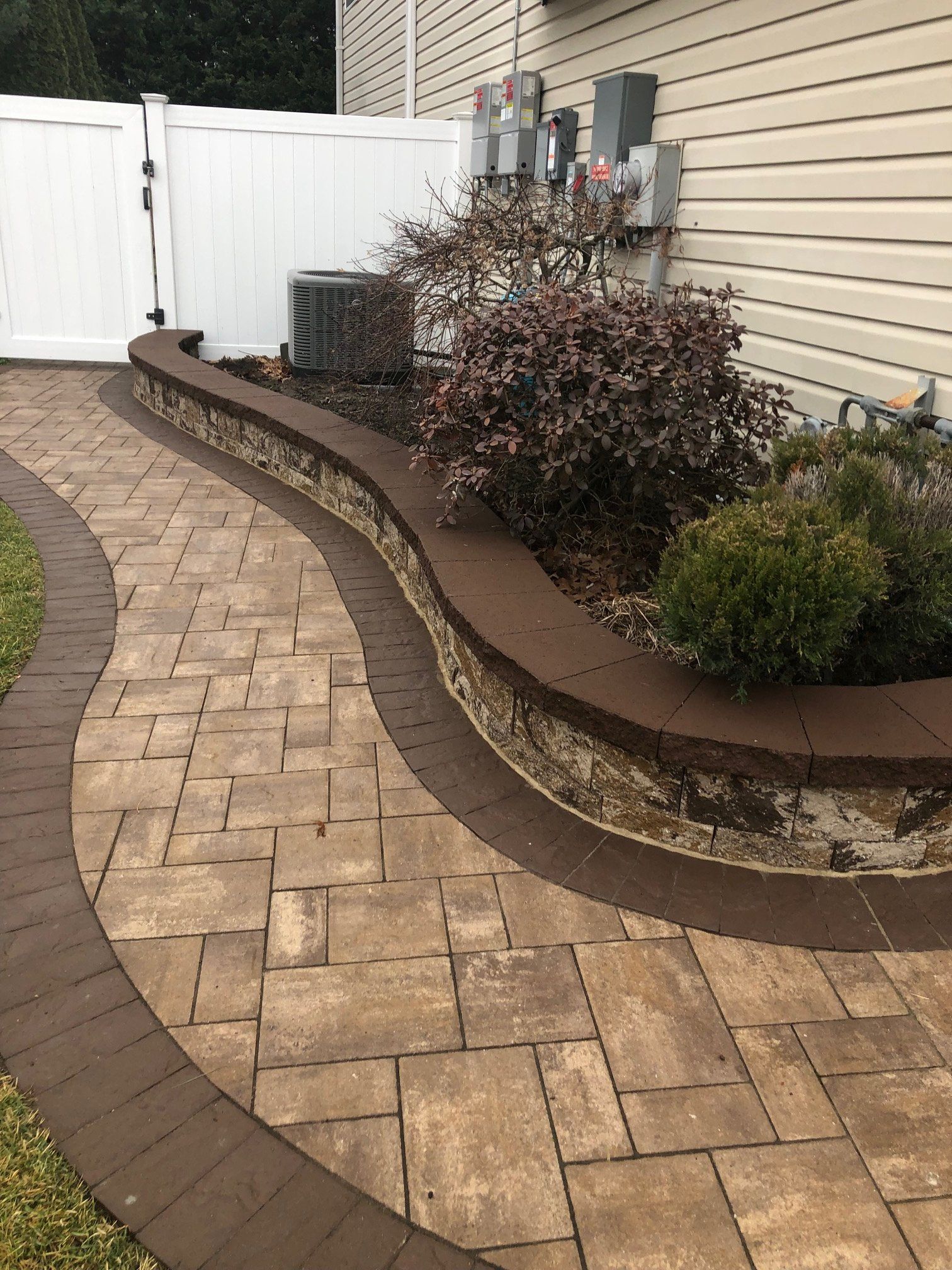 Curving brick pathway with brown border and raised garden bed along a house.