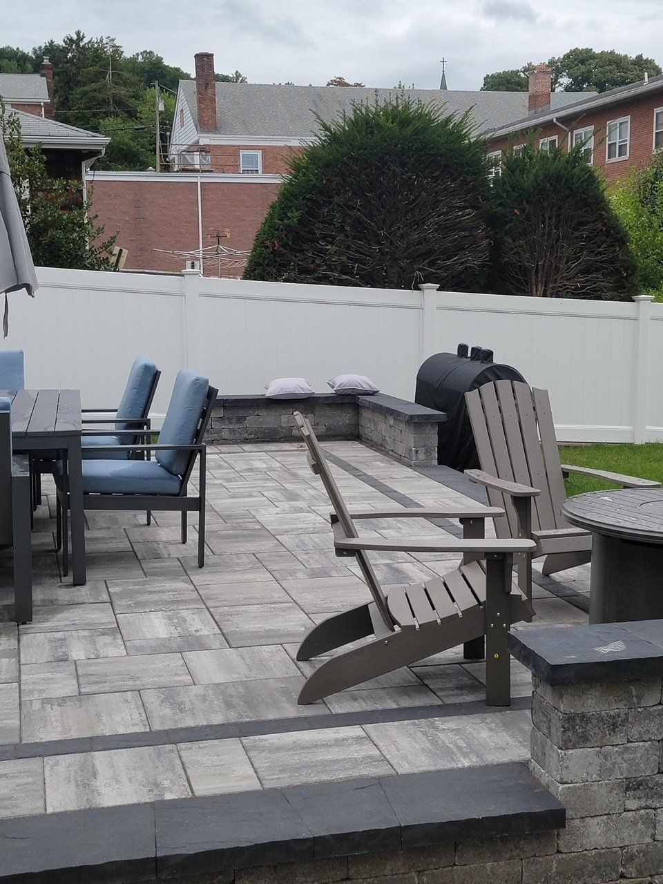 Patio with Adirondack chairs, dining set, and a grill against a white fence and brick buildings.