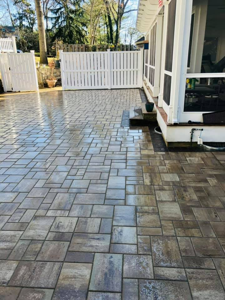 Brick patio with a white fence and house. It is wet, and a sunny day.
