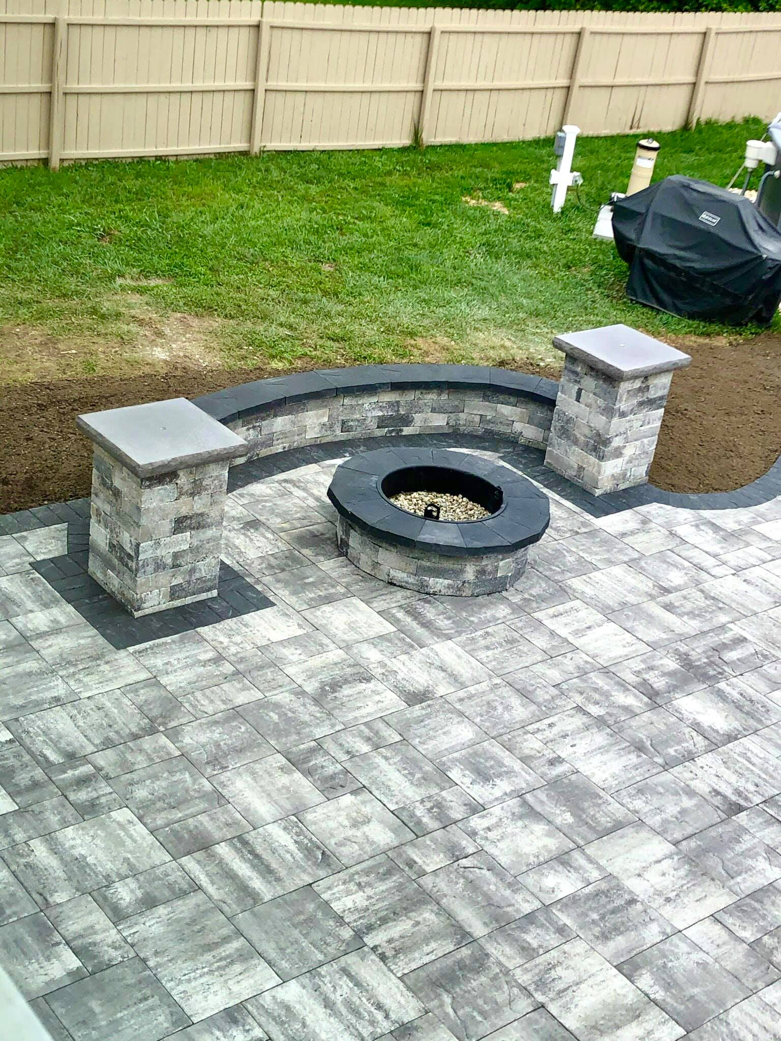 Patio with fire pit and seating, pavers in gray tones, grassy background.