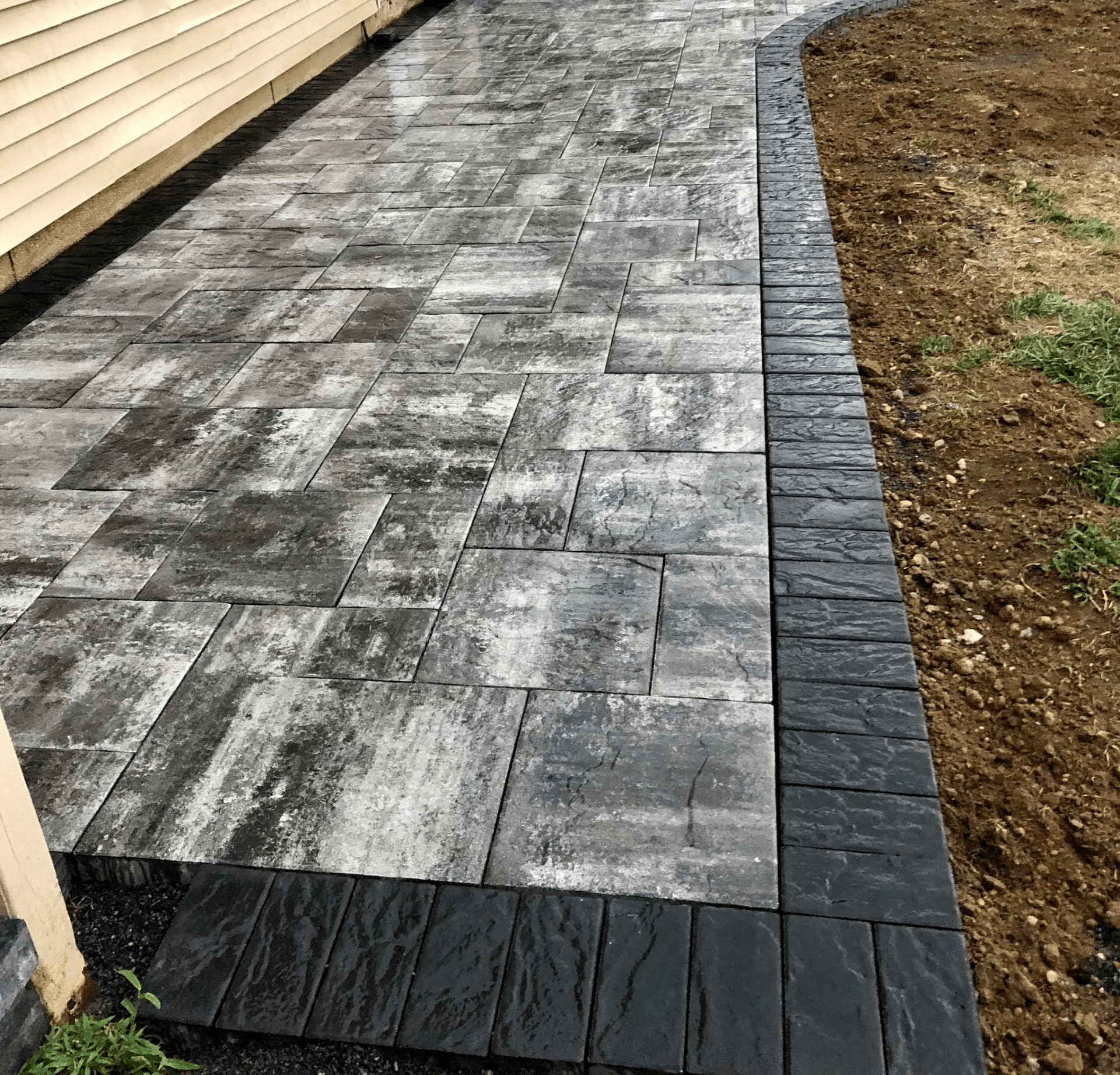 Gray and black paved walkway alongside a building and a grassy area.