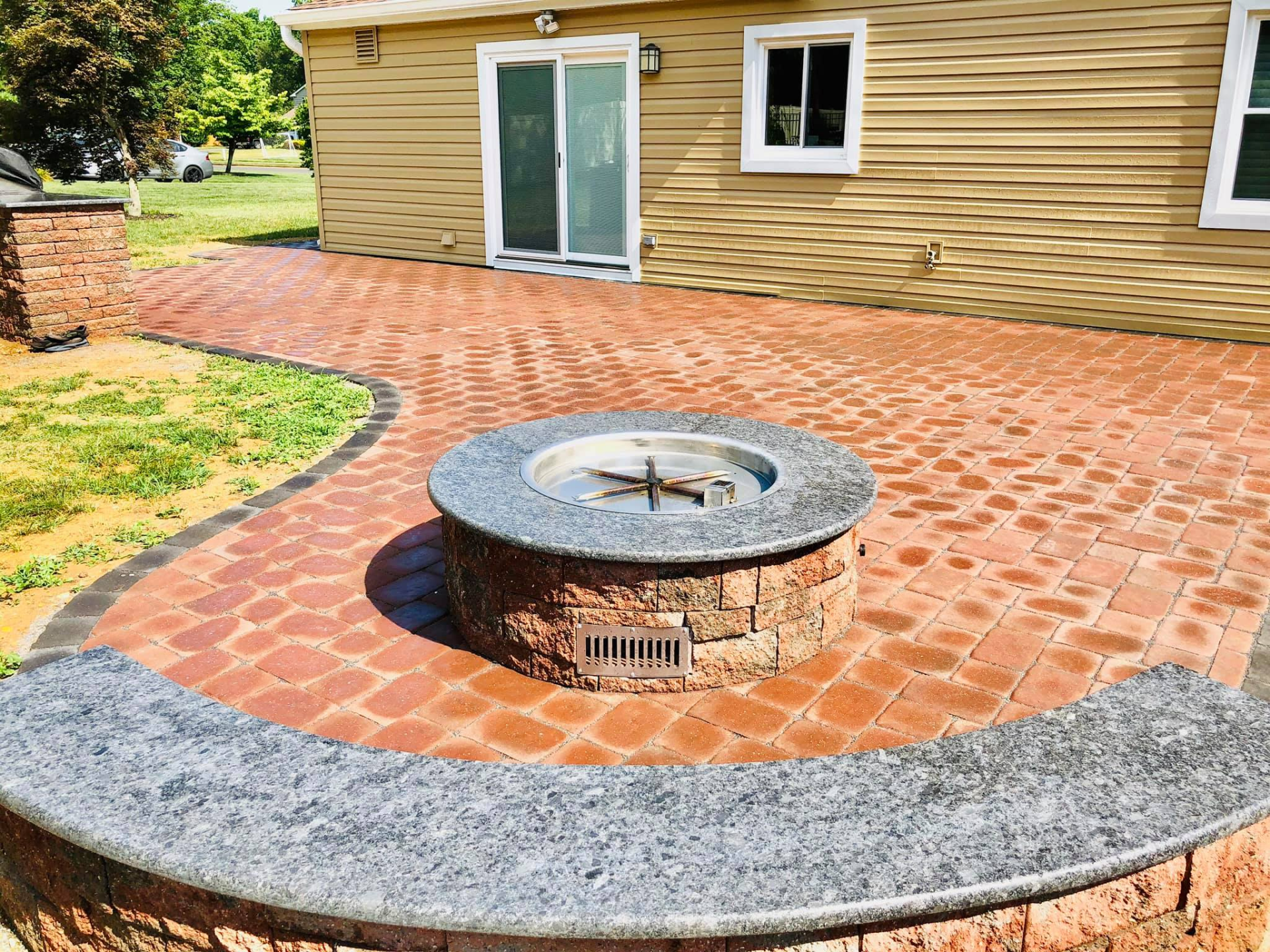 Brick patio with a fire pit and curved stone wall in front of a house.