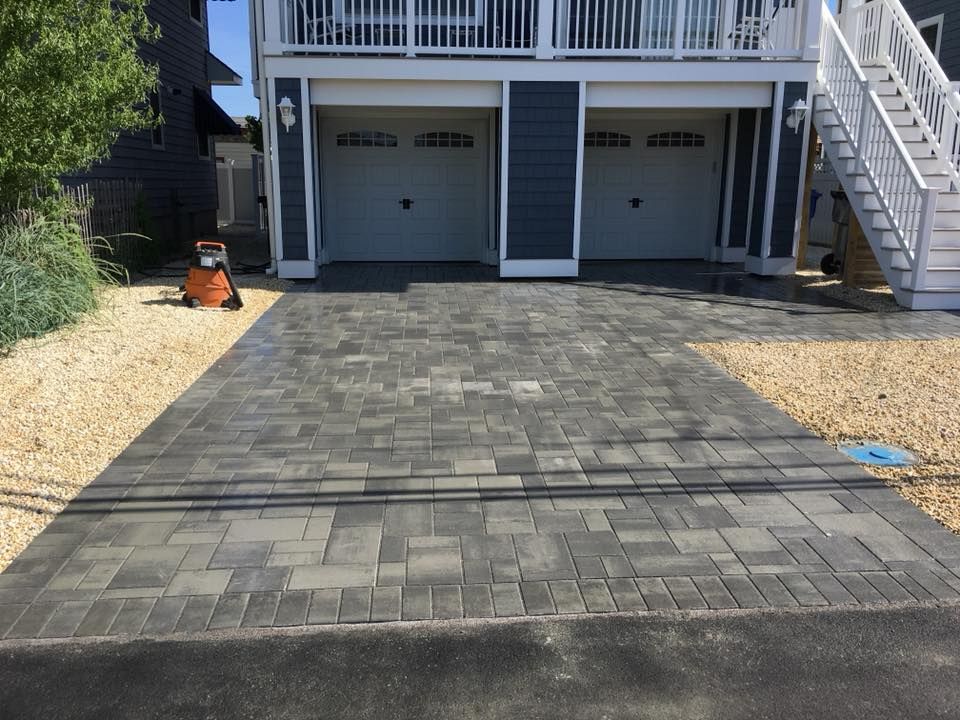 Brick paved driveway leading to a garage with two doors under a house with a staircase.