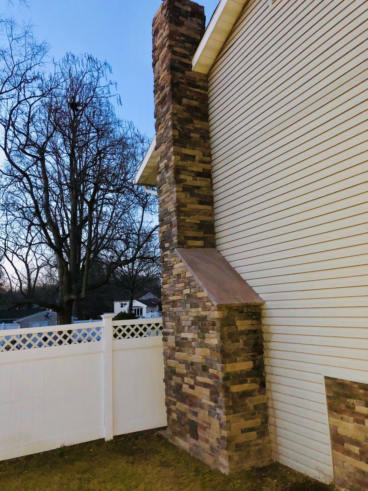 Tall stone chimney on a house with beige siding, next to a white fence and a leafless tree.