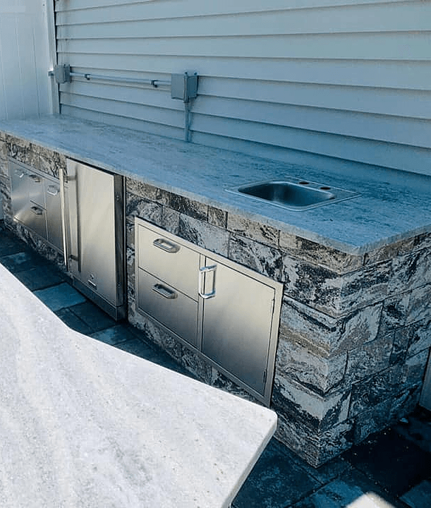 Outdoor kitchen with stone facade, stainless steel appliances, granite countertop, and sink against a white clapboard wall.