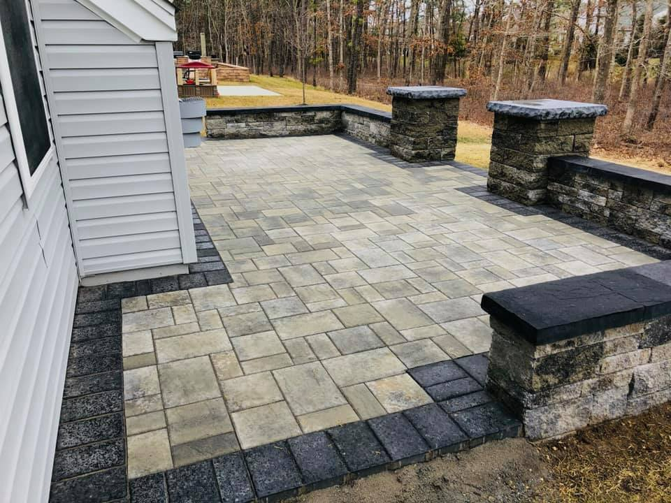 Stone patio with retaining walls.  Gray and black pavers border the patio near a white house.