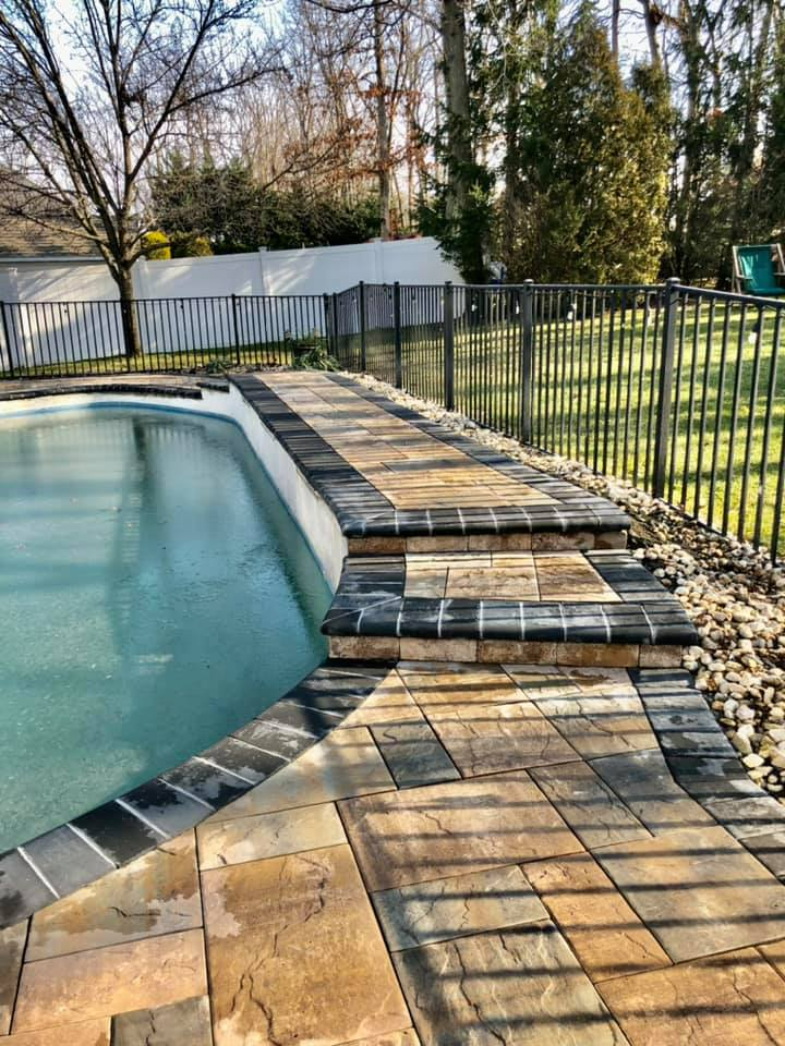 Poolside with steps and paving stones, enclosed by a black metal fence and trees.