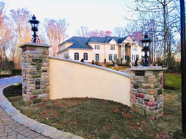 Stone pillars with lamps frame a tan wall and a large house with columns.