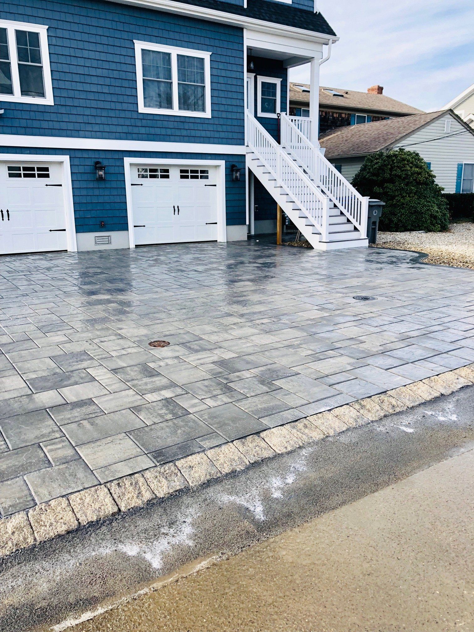Blue house with white garage doors and stairs, brick paved driveway, wet pavement.