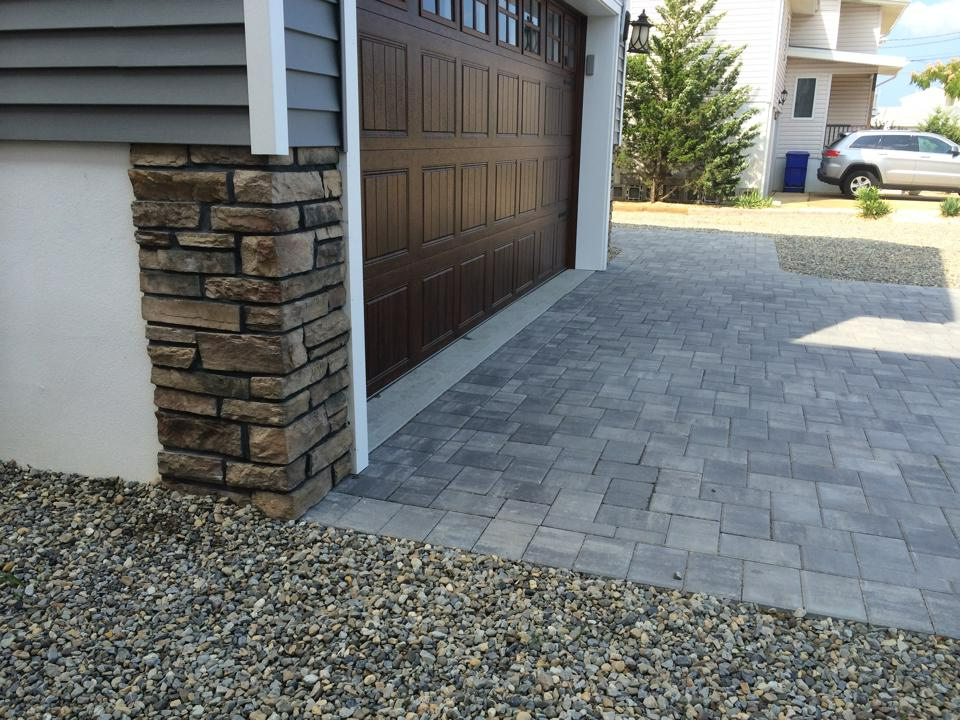 Garage exterior with stone column, brown garage door, and paved driveway. Gravel bed.