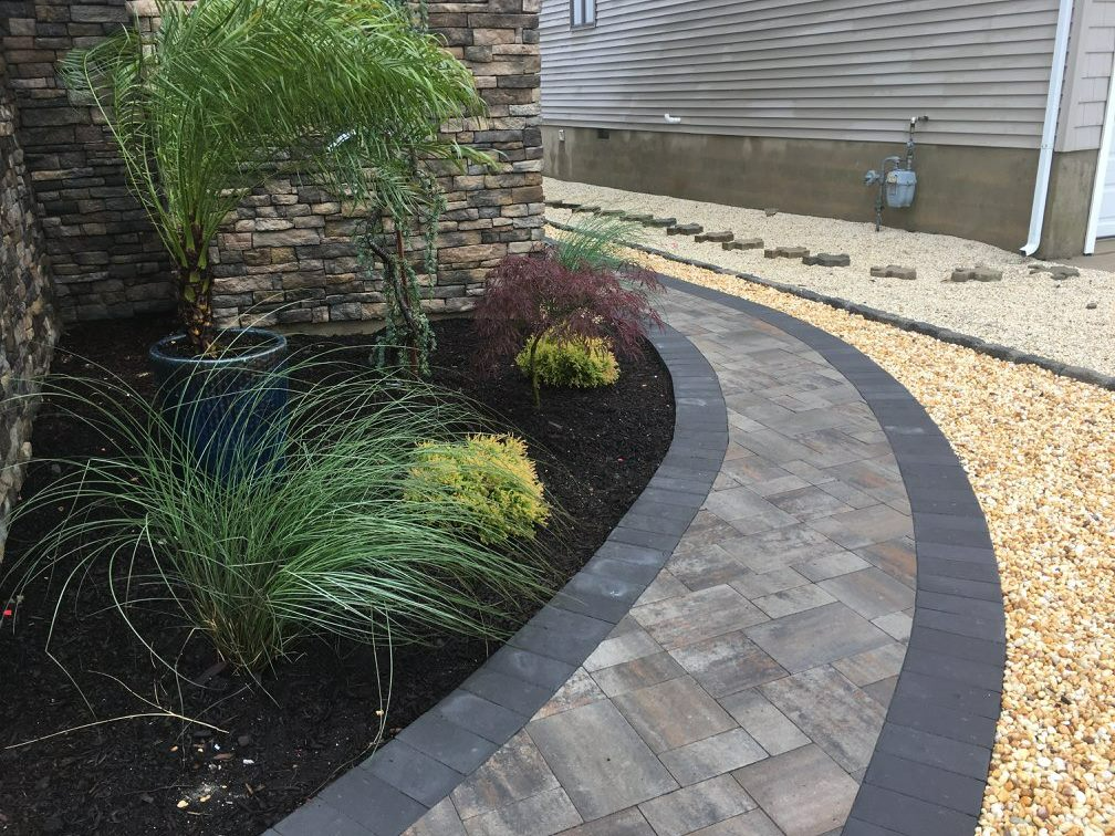 A landscaped walkway with pavers and dark edging curves alongside a house with a stone accent wall.