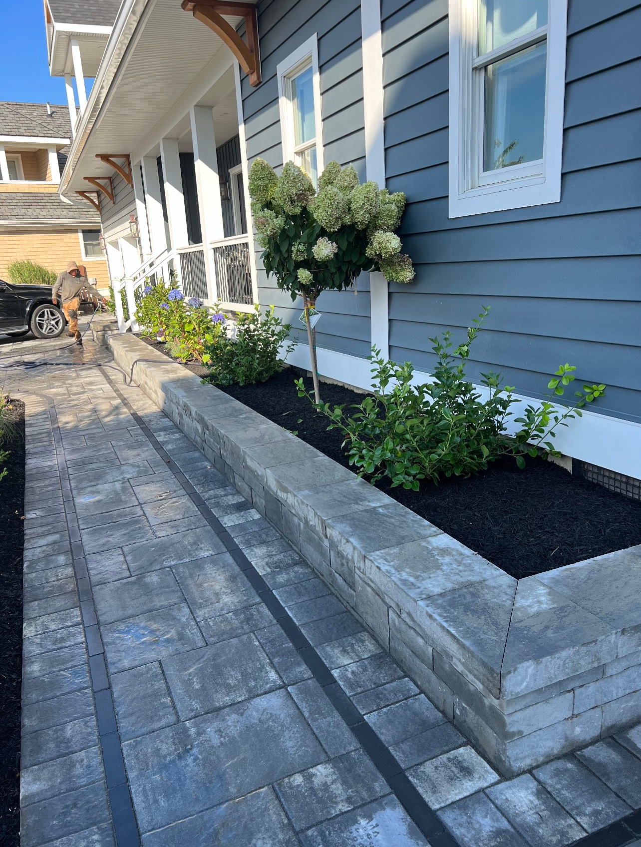 Gray brick walkway alongside a blue house with a raised garden bed.