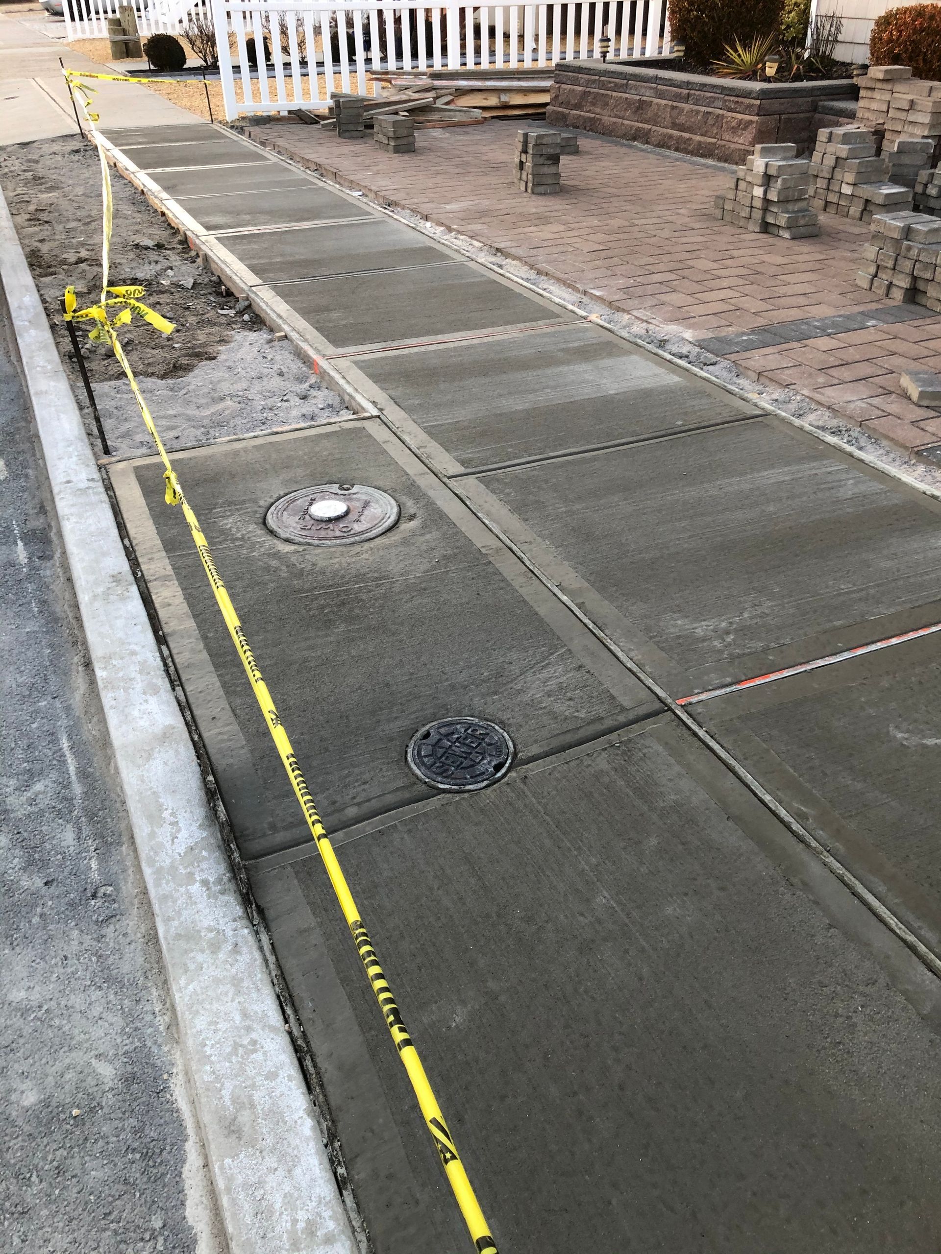 Newly poured concrete sidewalk with utility covers, cordoned off with caution tape.