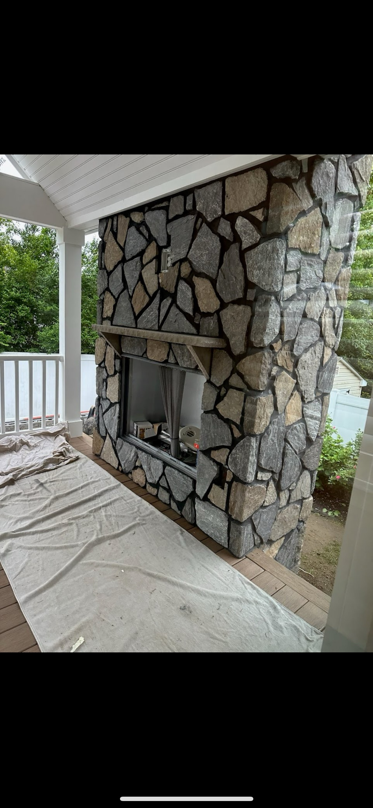 Outdoor stone fireplace on a porch with white railing. Fireplace has a wooden mantle and an exposed firebox.