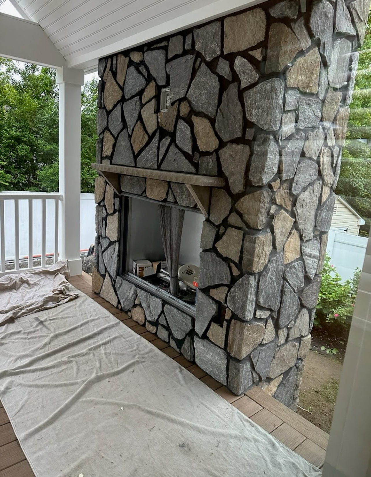 Stone fireplace on a porch; gray, beige stone with black grout. White trim and railing. Protective cloth on floor.