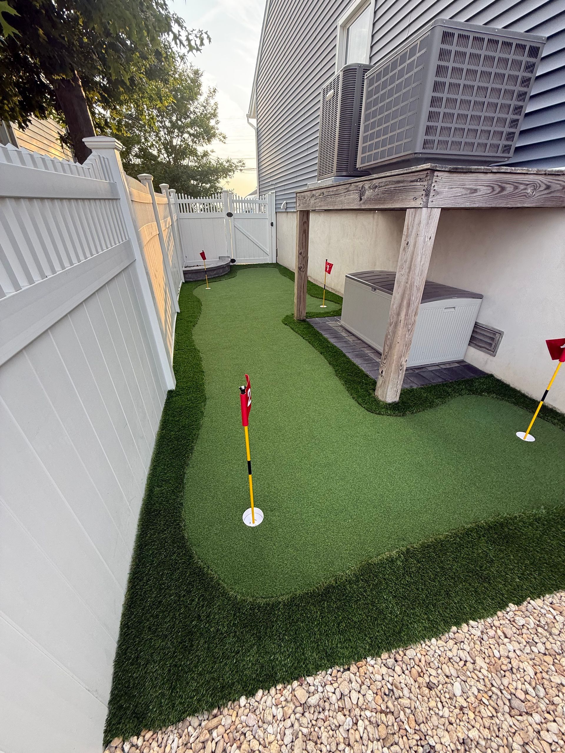 A backyard putting green with white fences. Two red flags mark the holes. A gray air conditioning unit sits in the corner.