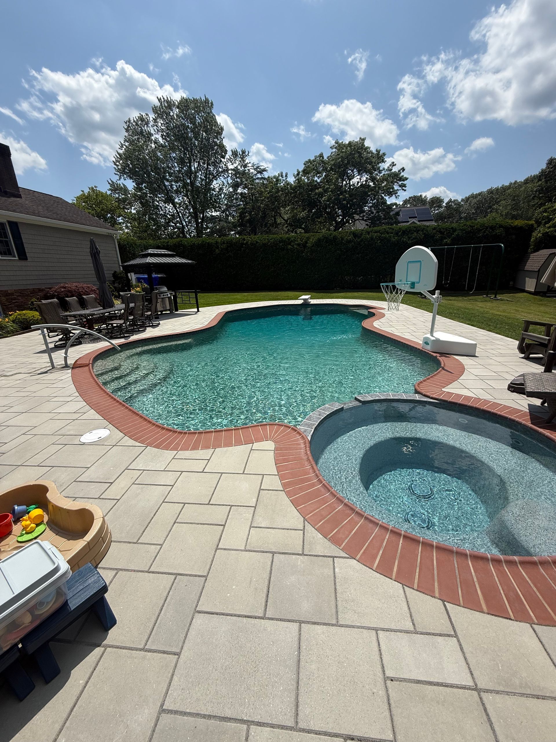 Backyard pool and hot tub on a sunny day. Paver patio, green lawn, trees, and blue sky in the background.