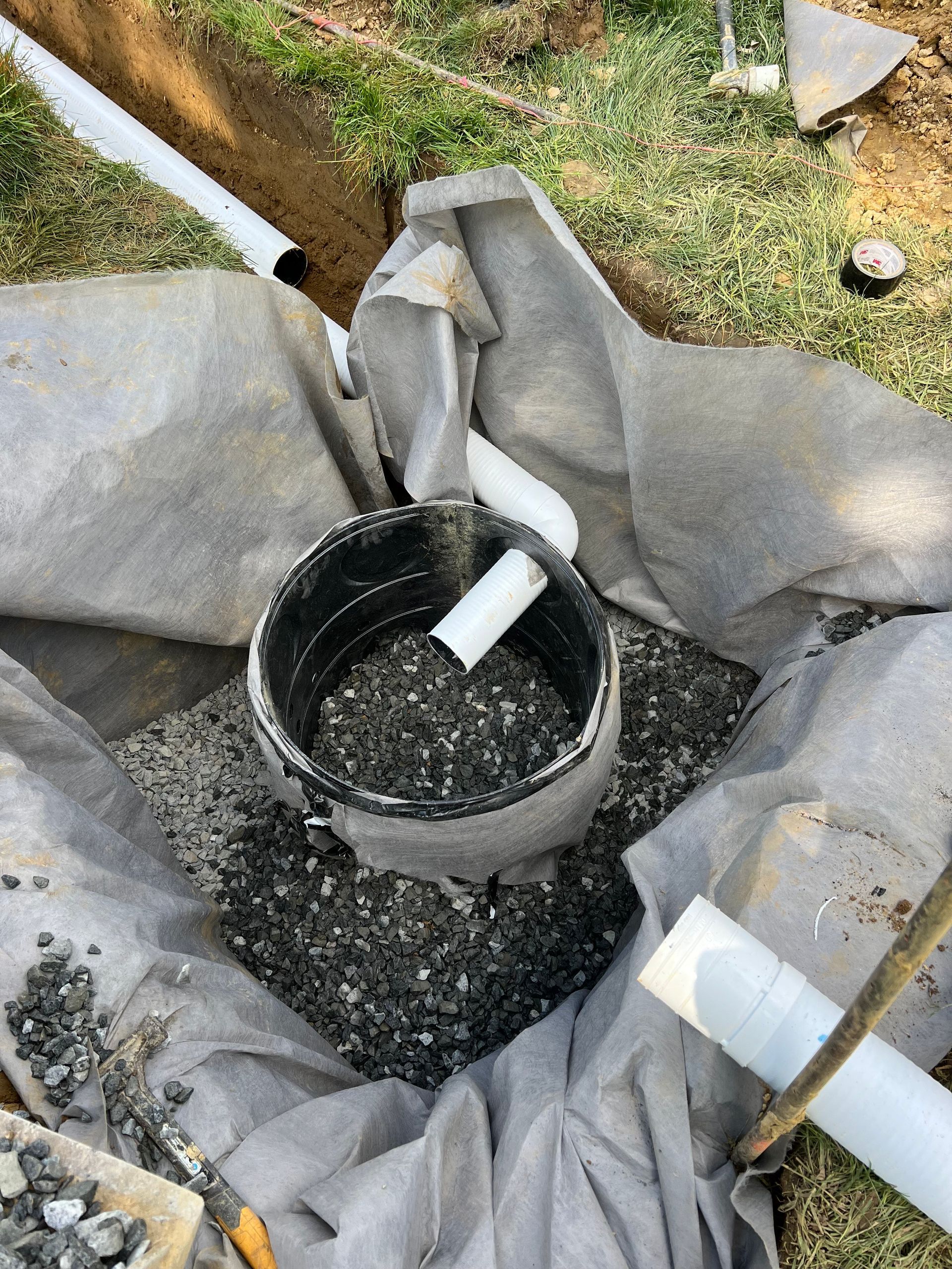 A partially buried black plastic catch basin surrounded by gravel and wrapped in fabric. White pipes are connected.
