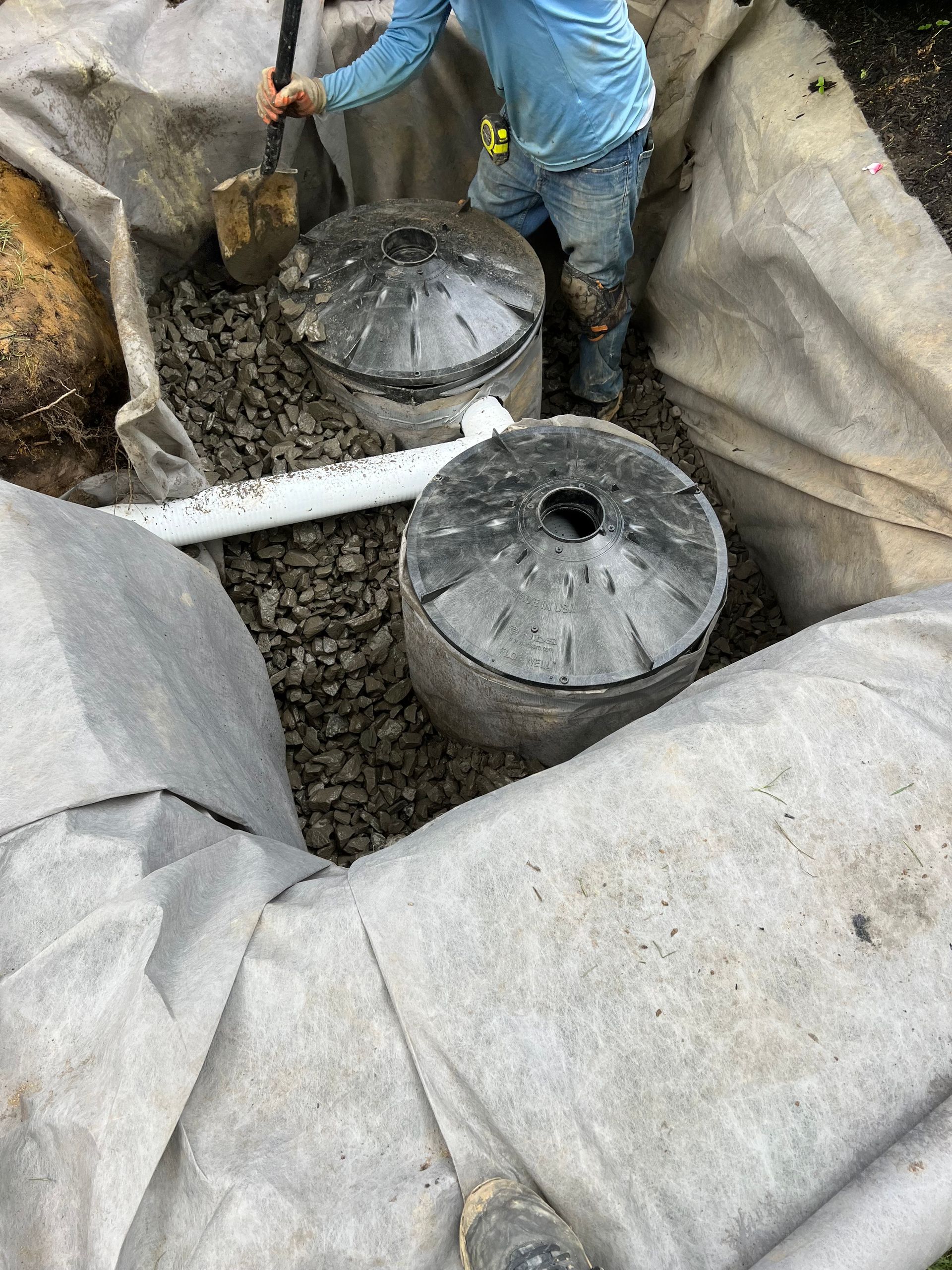 Person installing two large, round, gray tanks in a gravel-lined trench, covered in fabric.
