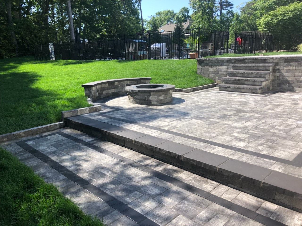 Stone patio with fire pit, built-in seating, steps, and green lawn in a backyard setting.
