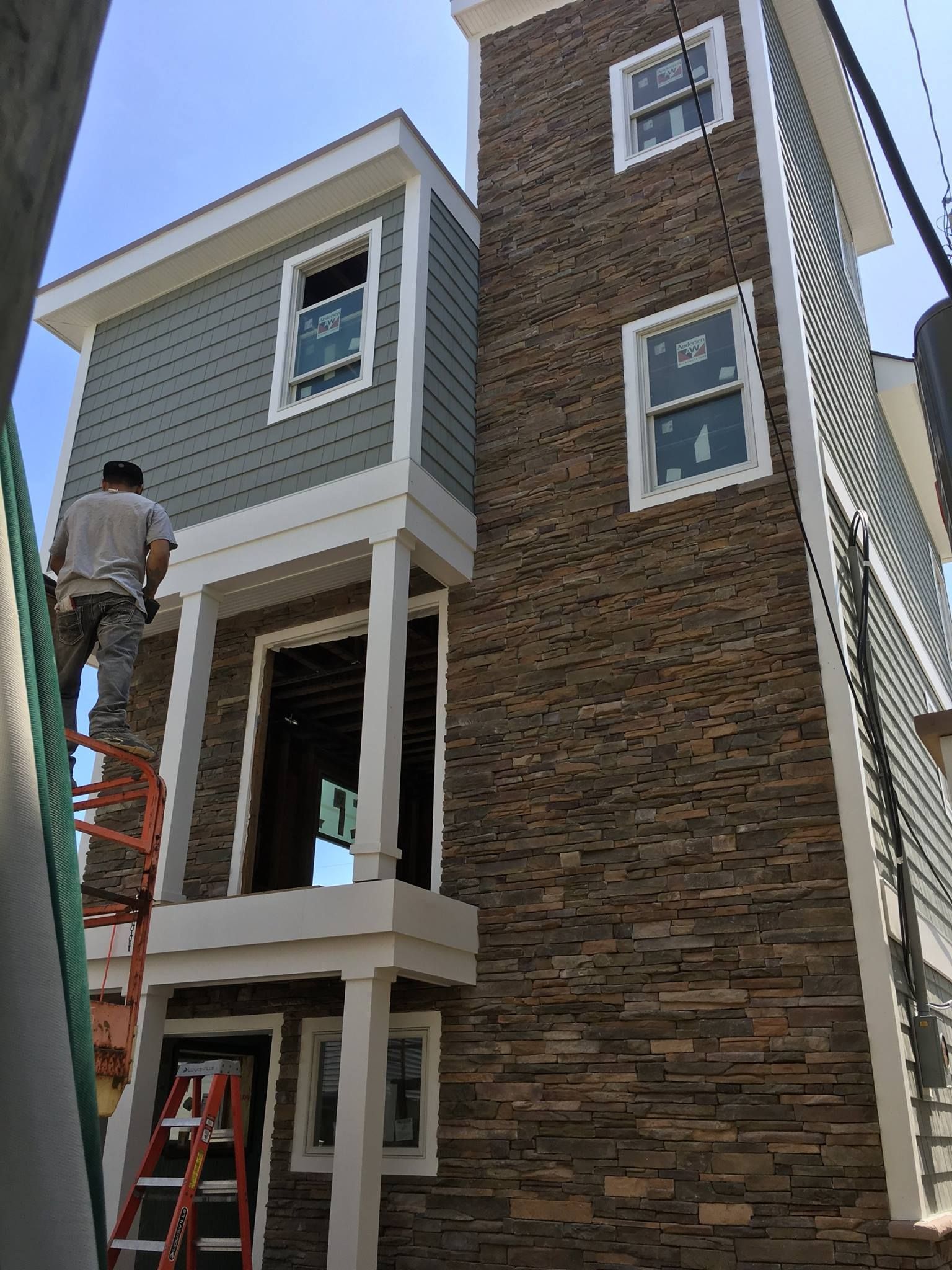 A worker on a ladder, installing exterior siding on a multi-story house with stone and gray siding.