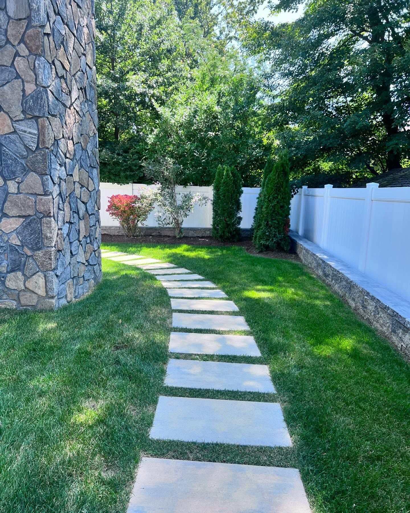 Stone path through a grassy yard alongside a stone wall and white fence.