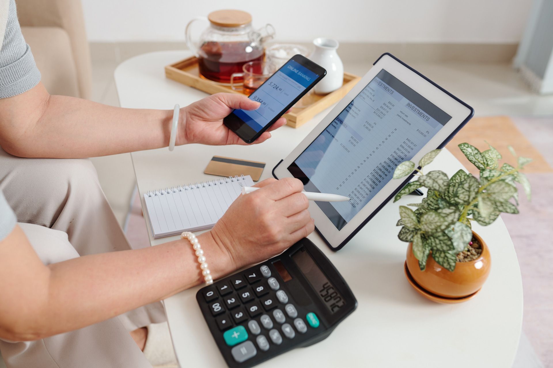A woman is sitting at a table using a tablet and a cell phone.