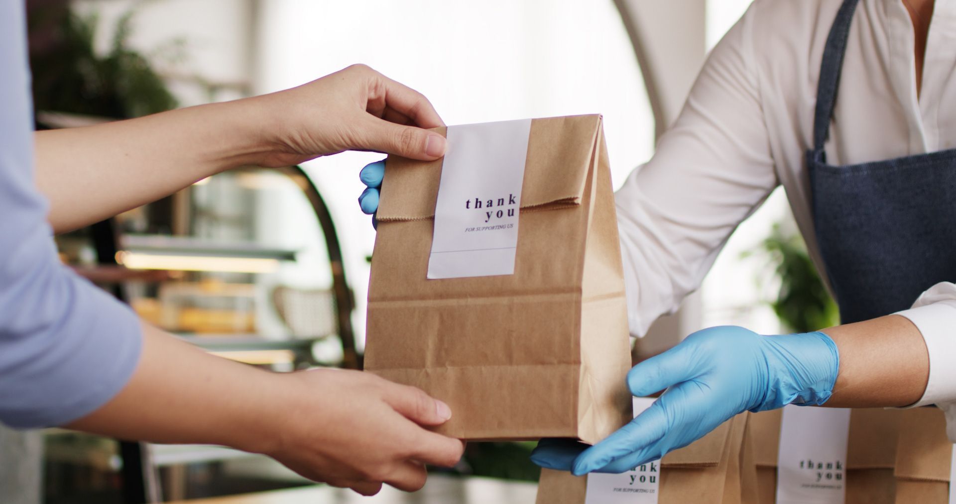 A person wearing blue gloves is handing a brown paper bag to another person.