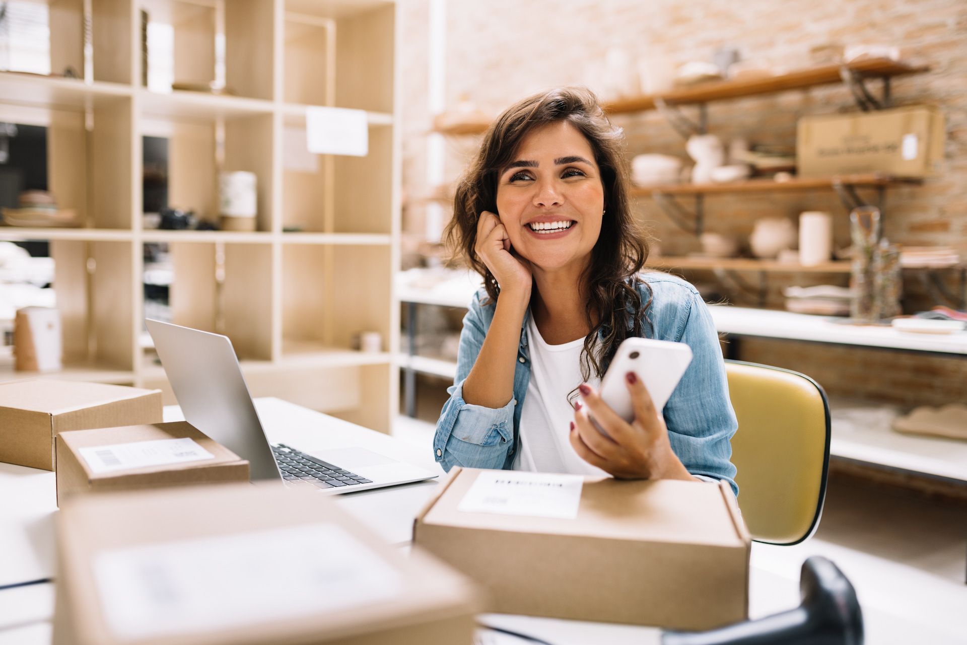 A woman is sitting at a desk holding a cell phone and a laptop.
