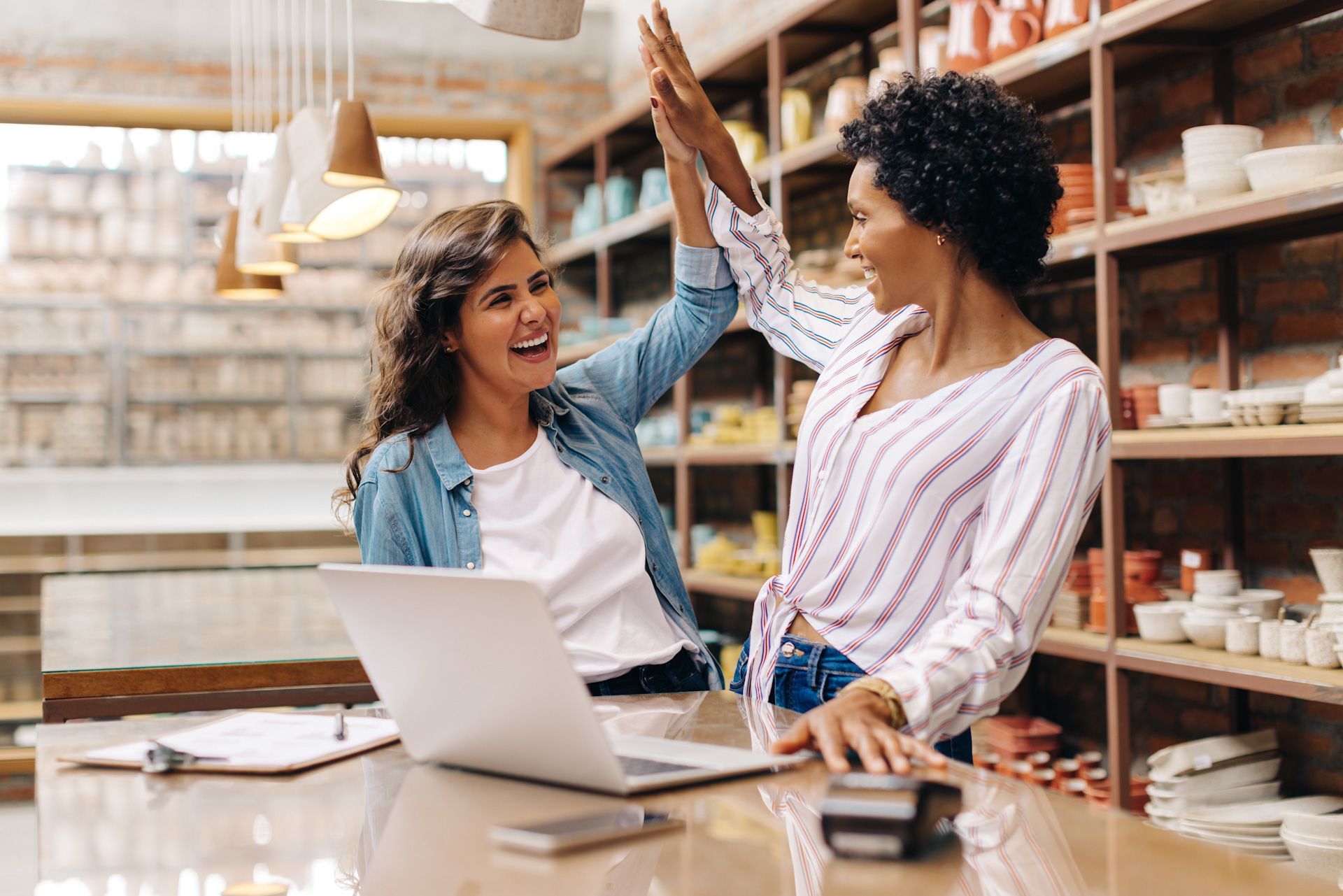 Two women are giving each other a high five while sitting at a table with a laptop.