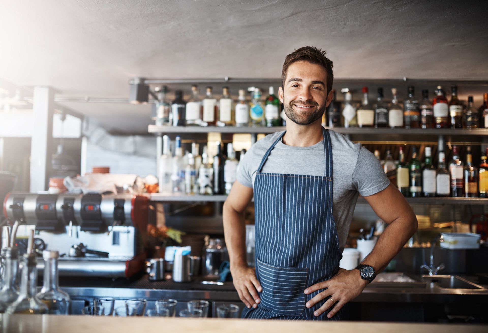 A man in an apron is standing in a restaurant with his hands on his hips.